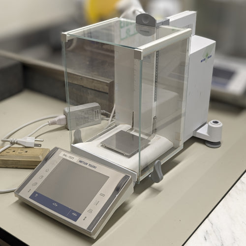 Mettler Toledo XP504 analytical balance ka front angle view, jisme glass draft shield, weighing pan, digital touch screen control panel, aur power cables saaf dikhai de rahe hain.