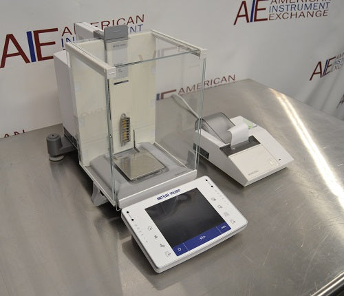Angled perspective view of a Mettler Toledo XPE205 analytical balance resting on a stainless steel surface, featuring its protective glass draft shield, separate touchscreen control terminal, weighing pan, and attached laboratory printer