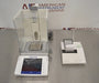 Front high-angle view of a Mettler Toledo XPE205 analytical balance on a stainless steel counter, featuring its glass draft shield, separate touchscreen terminal, weighing pan, and a connected laboratory printer