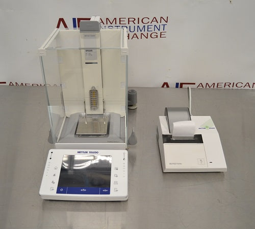 Top-down view of a Mettler Toledo XPE205 analytical balance resting on a stainless steel work surface, featuring its glass draft shield, internal weighing pan, front touchscreen terminal, and connected lab printer