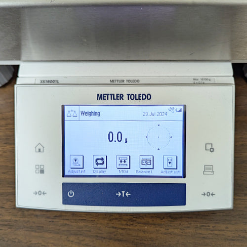 Close-up view of the Mettler Toledo XS16001L digital precision balance control terminal, featuring an active LCD screen showing a zero weight reading and touchscreen menu options, flanked by tactile operational buttons