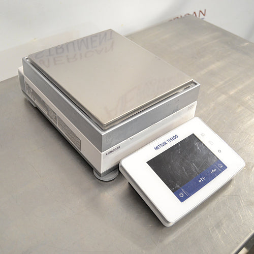Right angled perspective view of a Mettler Toledo XSR6002S Excellence precision toploading balance on a stainless steel surface, highlighting the rectangular weighing pan, grey and white main housing, and the detached front digital touchscreen terminal
