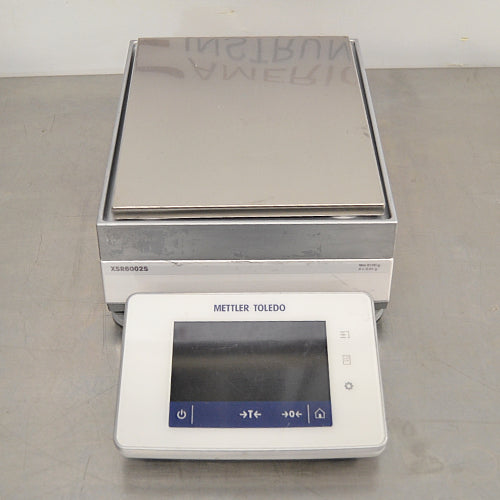 Front eye-level view of a Mettler Toledo XSR6002S Excellence precision toploading balance on a stainless steel surface, featuring its rectangular stainless steel weighing pan, grey and white housing, and a front digital touchscreen terminal with blue function icons