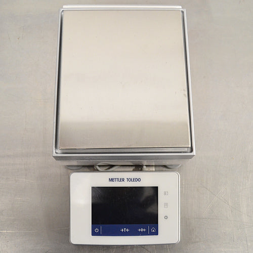 Top-down eye-level view of a Mettler Toledo XSR6002S Excellence precision toploading balance on a stainless steel surface, highlighting the flat rectangular stainless steel weighing pan and the front digital touchscreen terminal