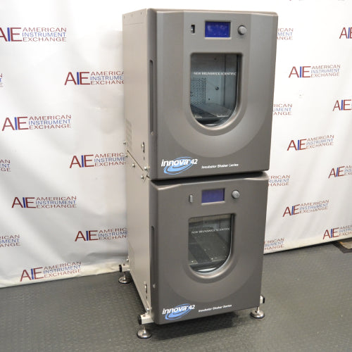 Left three-quarter angle product view of a New Brunswick Innova 42R stacked refrigerated incubator shaker system, showcasing the dual stacked grey chassis units, curved doors with central viewing windows, digital displays, and adjustable leveling feet