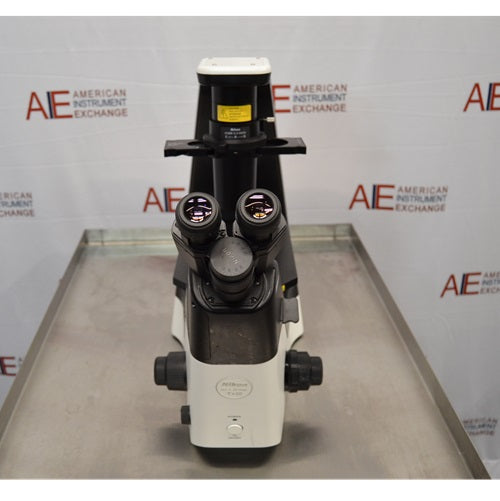 Direct front-on view of a Nikon Eclipse Ts2 inverted microscope resting on a stainless steel surface, highlighting the symmetrical layout of the lower housing, power button, coaxial focus knobs, binocular eyepieces, and overhead condenser tower