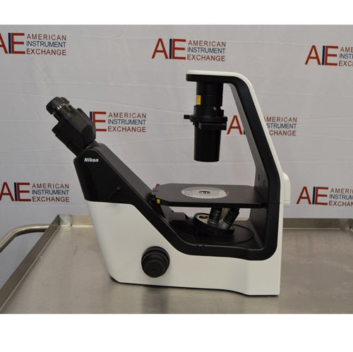 Left profile view of a Nikon Eclipse Ts2 inverted microscope on a stainless steel surface, featuring its black-and-white ergonomic frame, stage, coarse and fine focus controls, overhead condenser column, and the left-facing binocular viewing head