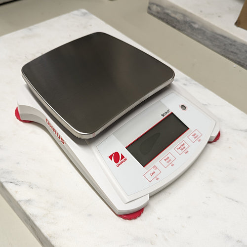 Front three-quarter product view of an Ohaus Scout SPX2201 portable balance, showcasing its stainless steel pan, white housing, red brand logo, and angled digital display panel