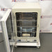 Studio angle product view of a Panasonic Cell-IQ Series MCO-170AICUVL-PA CO2 cell culture incubator with the outer door swung open against a branded backdrop, highlighting its off-white housing, open internal chamber with perforated stainless steel shelves, a lower water reservoir pan, and wheeled platform base