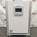 Studio front product view of a PHCBi Cell-IQ Series CO2 laboratory incubator against a branded backdrop, highlighting its large white outer cabinet, vertical silver side pillars, central door-mounted digital touchscreen control panel with PHCBi branding, and mobile wheeled base platform