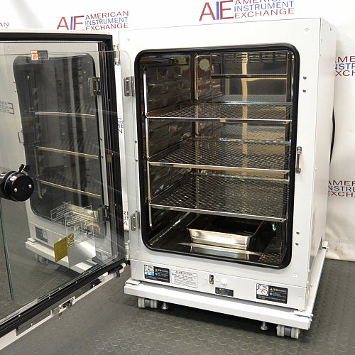 Studio angle product view of a PHCBi Cell-IQ Series CO2 laboratory incubator with the outer door swung open against a branded backdrop, highlighting its white housing, heavy door hinges, open inner chamber with multiple stainless steel wire shelves, a lower water pan, and wheeled base platform