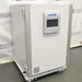 Studio three-quarter angle product view of a PHCBi Cell-IQ Series CO2 laboratory incubator against a branded backdrop, highlighting its large white outer cabinet, heavy-duty door hinges on the side, vertical silver pillars, central touchscreen display, and wheeled base platform
