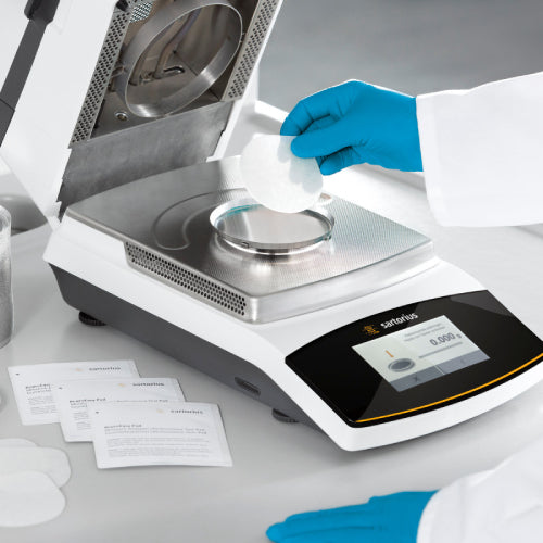 Left angle action view of a Sartorius MA160 moisture analyzer, with a gloved hand placing a sample pad onto the weighing pan inside the open heating chamber, alongside accessory packets and a touch control display