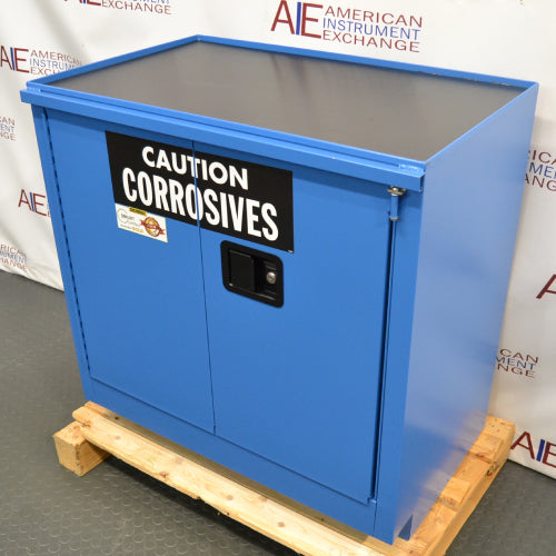 Left three-quarter angle product view of an unused Securall 24-gallon corrosive storage cabinet, focusing on the blue side panel, warning label, and secure door latch mechanism on a wood pallet