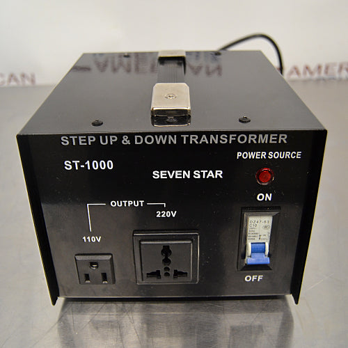 Straight-on front view of a Seven Star ST-1000 step up and down voltage transformer, featuring the 110V and 220V output sockets, blue toggle power switch, power source indicator light, and top handle on a stainless steel surface