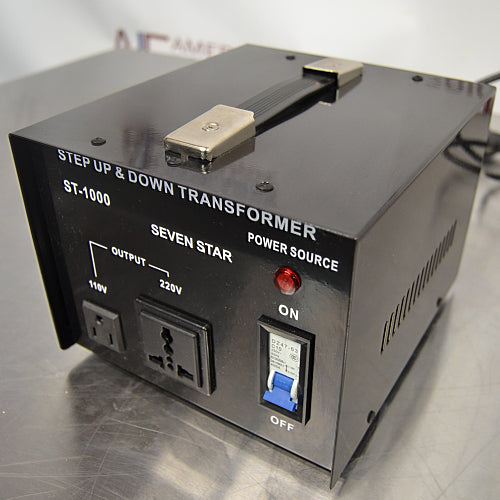 Three-quarter angle product view of a Seven Star ST-1000 step up and down voltage transformer, highlighting its black metallic chassis, top carry handle, 110V and 220V output sockets, power switch, and indicator light on a lab bench