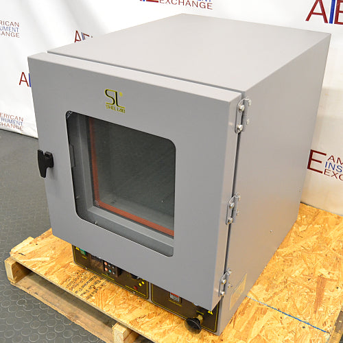 Right angled perspective view of a Shel Lab 1445 vacuum oven resting on a wooden pallet, highlighting its grey housing, glass viewing door with a latch handle, exterior door hinges, and lower control panel