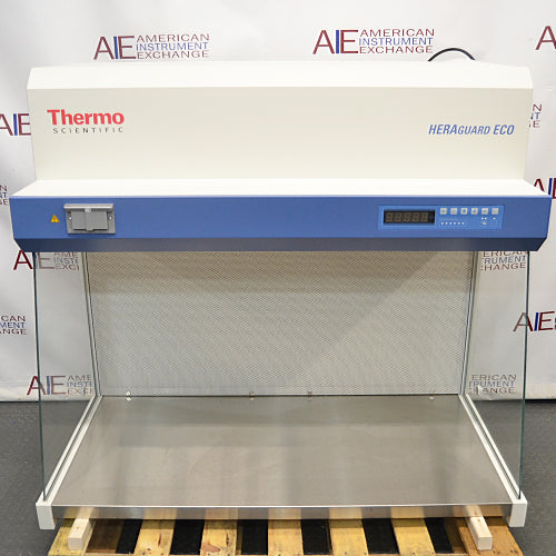 Front product view of a Thermo Scientific HeraGuard ECO 1.2 laminar flow clean bench, showcasing its white and blue housing, digital control panel, stainless steel work surface, and glass side walls on a wooden pallet