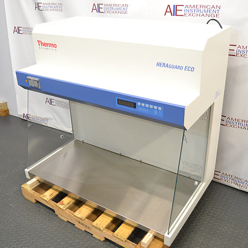 High-angle three-quarter product view of a Thermo Scientific HeraGuard ECO 1.2 laminar flow clean bench, showcasing its white housing, blue control console, stainless steel work deck, and glass panels on a wooden pallet