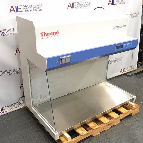 Right three-quarter angle product view of a Thermo Scientific HeraGuard ECO 1.2 laminar flow clean bench, featuring its white and blue exterior, digital control panel, stainless steel work deck, and glass side panels on a wooden pallet