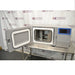 Front eye-level view of a Thermo Scientific CryoMed controlled rate freezer resting on a stainless steel surface with its heavy-duty chamber door open, revealing the internal sample racks and chamber insulation