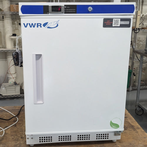 Front product view of a VWR HCUCBI0404 undercounter laboratory refrigerator, featuring a white solid door with vertical handle, blue and black control panel console at the top with digital display, keyed lock, and lower ventilation grille on a wooden surface